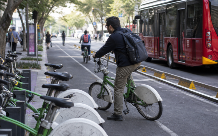 Persona usando una bicicleta pública ECOBICI junto a una cicloestación y una avenida de Ciudad de México.