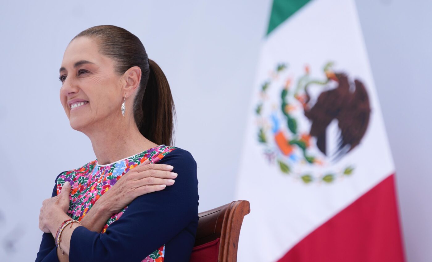 Claudia Sheinbaum sonríe sentada junto a la bandera de México durante el acto.